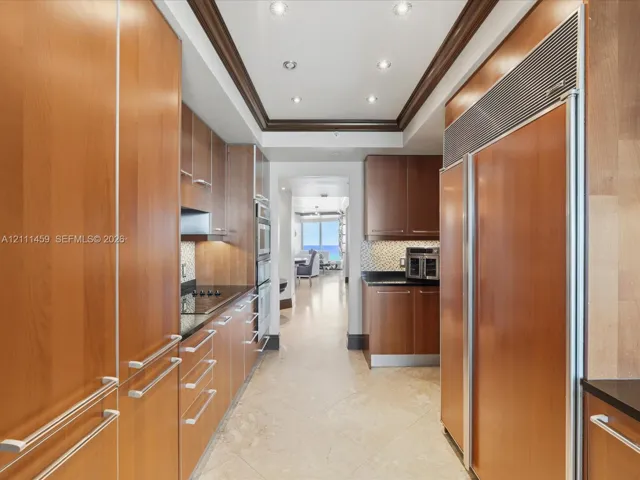 Galley kitchen featuring custom flat-panel wood cabinetry, integrated stainless steel refrigerator, black stone countertops, tile flooring, and recessed ceiling lighting with crown molding