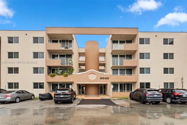 Multi-story exterior featuring a neutral stucco facade, private recessed balconies with metal railings, a central architectural tower, and dedicated paved surface parking