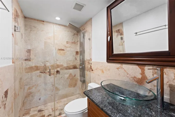 Walk-in shower with stone tile surround, glass panel enclosure, and integrated shower panel system, featuring a vanity with a dark stone countertop, glass vessel sink, and chrome faucet