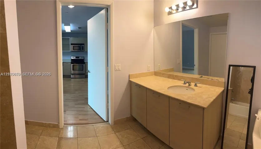 Bathroom featuring a light wood-finish vanity, stone-surfaced countertop with an integrated basin, wall-to-wall vanity mirror, four-bulb overhead vanity lighting, and ceramic tile flooring