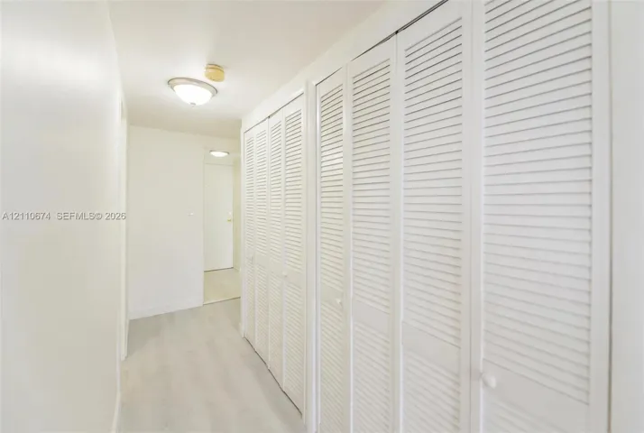 Full-length hallway featuring white louvered bifold closet doors, light-toned wood-finish flooring, and a flush-mount ceiling light fixture