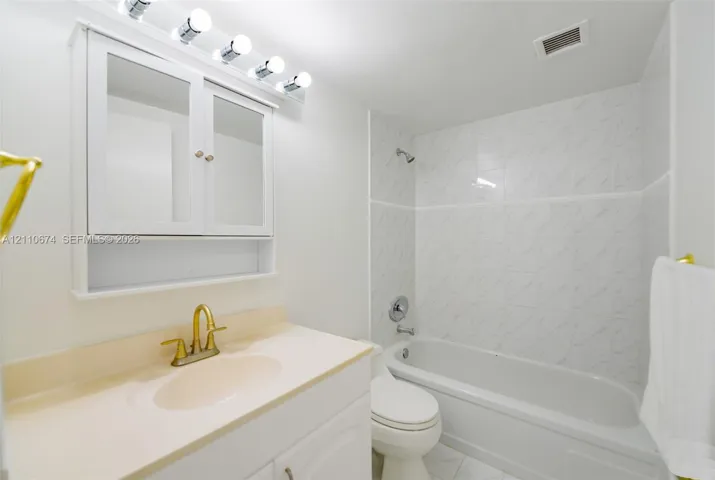 Bathroom featuring a vanity with integrated countertop and brass-finish faucet, white wall-mounted medicine cabinet with integrated lighting, and a shower-tub combination with patterned tile surround