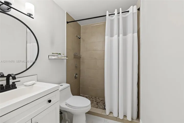 White vanity with integrated rectangular sink and matte black faucet, round wall-mounted mirror, matte black vanity lighting, shower enclosure with neutral-toned wall tiles, and stone-tiled shower flooring