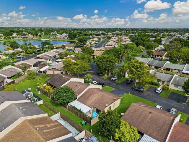 Aerial perspective of a residential community featuring shingle-roofed structures, expansive green space, mature trees, asphalt roadways, and a lake