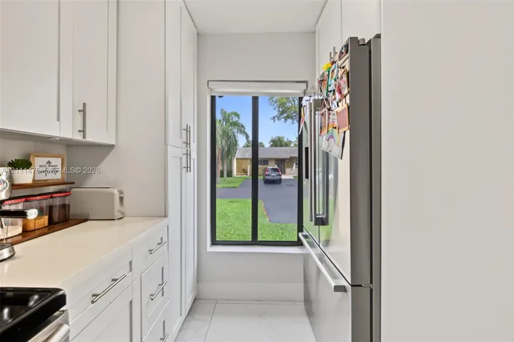 White shaker-style cabinetry, solid-surface countertops, floor-to-ceiling pantry storage, stainless steel refrigerator, and large-format tile flooring
