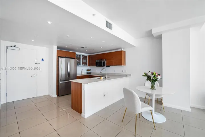 Kitchen featuring wood-finish cabinetry, stainless steel refrigerator, light-toned stone countertops, and recessed ceiling lighting with tile flooring