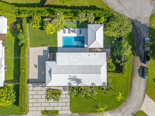Aerial view of property featuring a metal roof, rectangular swimming pool, expansive concrete paver driveway, manicured lawn, and mature perimeter hedging