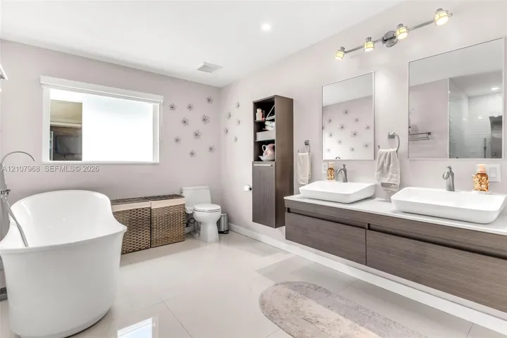 Floating vanity featuring wood-finish cabinetry, dual vessel sinks, and stone-surface countertop, complemented by a freestanding soaking tub, vertical built-in storage cabinet, and rectangular wall-mounted mirrors