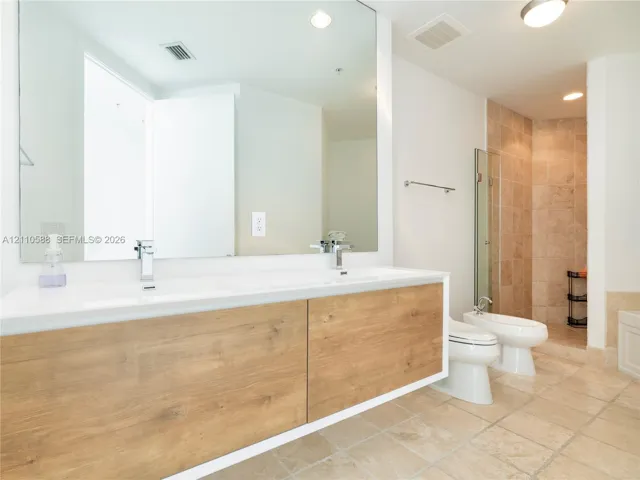 Floating dual-sink vanity with wood-finish cabinetry, expansive wall-mounted mirror, stone-tile flooring, and glass-enclosed shower