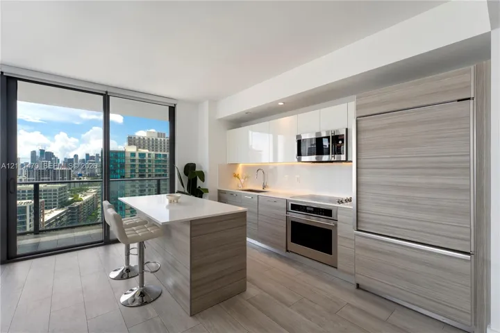 Contemporary kitchen featuring wood-finish cabinetry, white gloss upper cabinets, a solid-surface island, stainless steel appliances, and floor-to-ceiling glass sliding doors