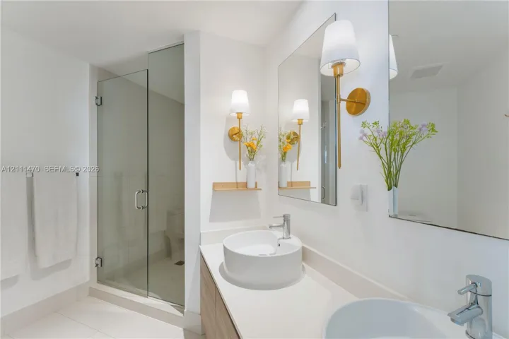 Bathroom featuring a dual-sink vanity with light-tone wood cabinetry, a quartz countertop, glass-enclosed shower, and gold-finish wall sconces