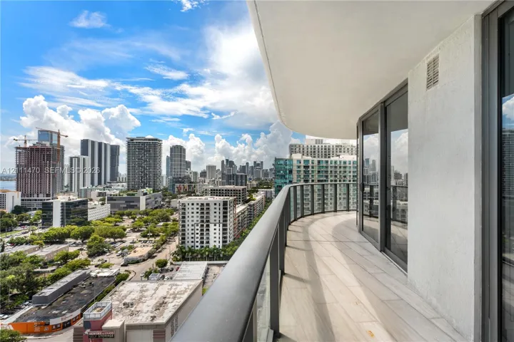 Curved balcony featuring light-toned tile flooring, glass railings with a dark metal frame, and sliding glass entry doors