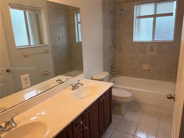 Double-sink vanity with a solid-surface countertop, dark wood-finish cabinetry, ceramic tile flooring, and a shower-tub combination featuring full-height wall tiling