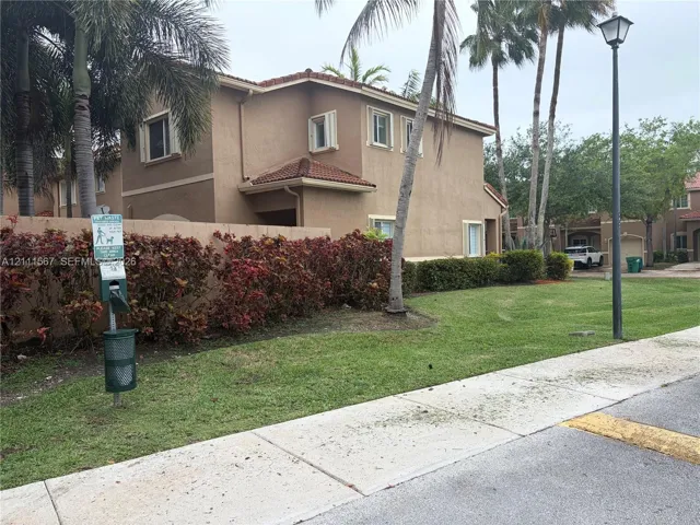 Two-story stucco exterior featuring a tile roof, mature palm trees, landscaped shrubbery, and a concrete sidewalk