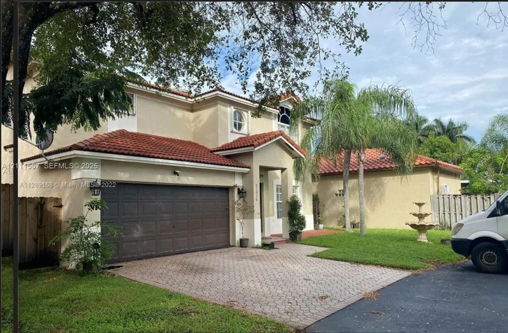 Stucco exterior residence featuring a multi-gabled tile roof, paver driveway, attached two-car garage, and tiered garden fountain
