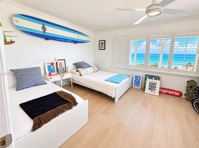 Light-toned wood-finish flooring, multi-pane windows with white plantation shutters, ceiling-mounted light fixture with integrated fan, and ocean scenery