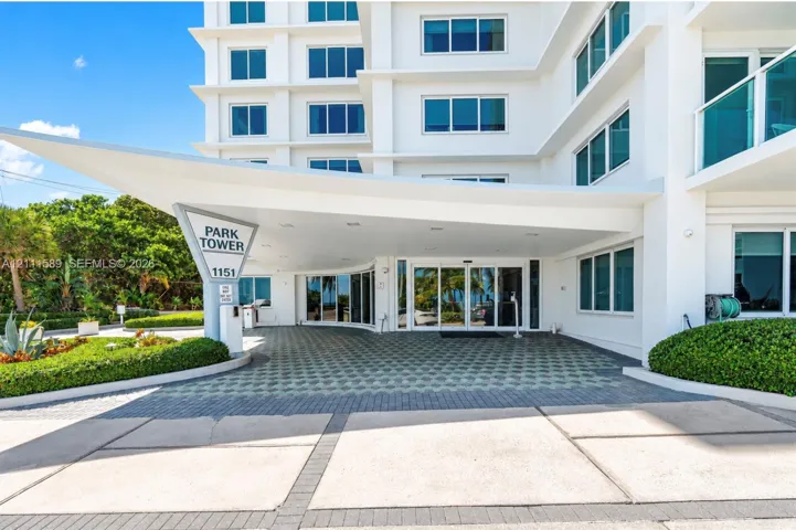 Modern high-rise exterior featuring a cantilevered covered entryway, glass sliding doors, concrete and paver drive, and manicured perimeter landscaping