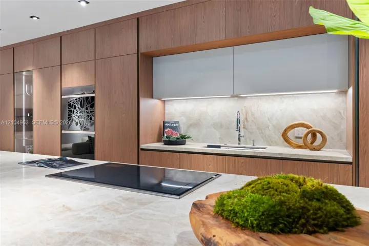 Modern kitchen featuring floor-to-ceiling wood-finish cabinetry, a large stone-surface island with an integrated induction cooktop, light-toned stone backsplash, and recessed ceiling lighting