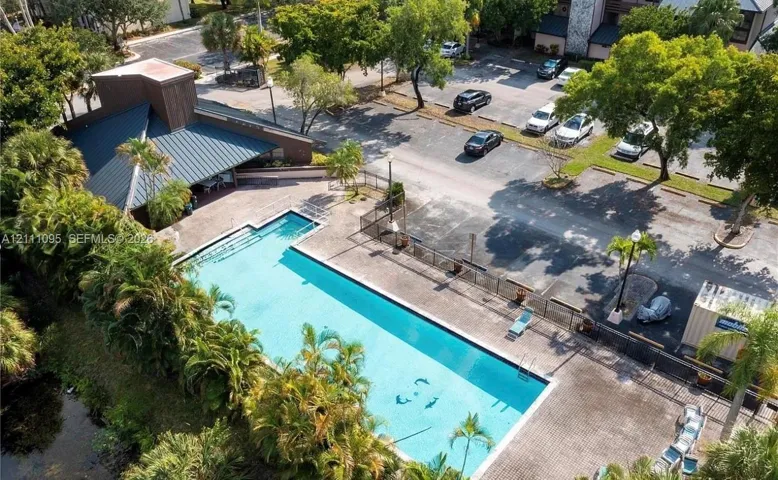 Rectangular swimming pool featuring a paved pool deck, metal perimeter fencing, covered pavilion, and mature tropical landscaping