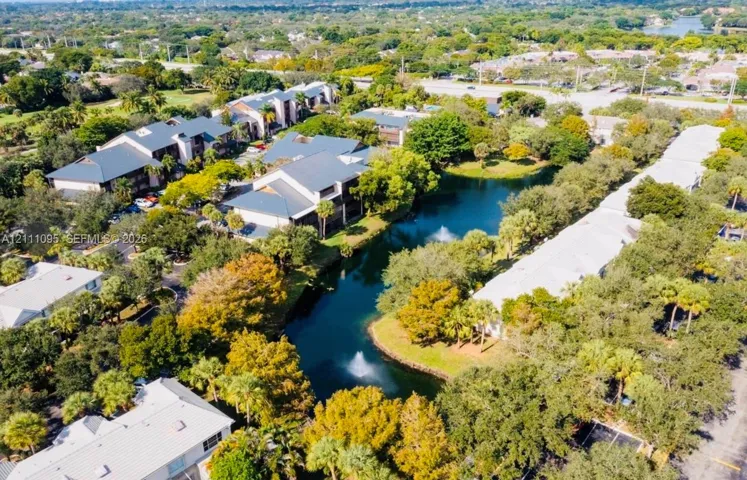 Aerial perspective featuring multi-story residential buildings, a central pond with a fountain feature, abundant mature canopy trees, and paved surface parking