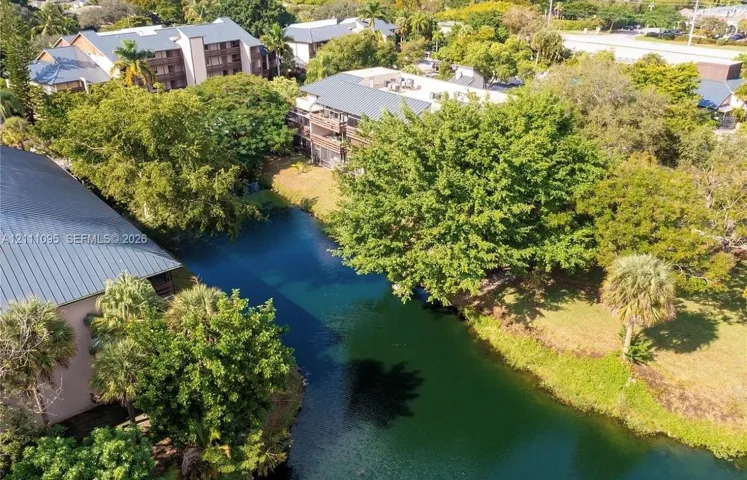 Aerial perspective featuring a multi-story building with a metal roof, situated alongside a water feature, accented by mature trees and palm foliage