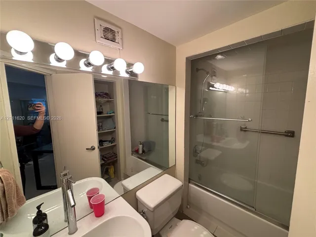 Bathroom featuring a single-sink vanity with a solid-surface countertop, a full-width wall mirror, a bathtub-shower combination with sliding glass doors and a grab bar, and built-in shelving