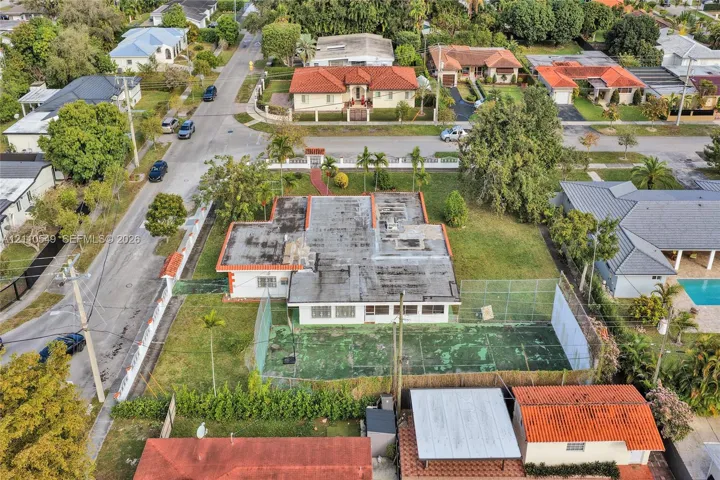 Aerial perspective of a corner lot property featuring a single-story residence, a private fenced-in sport court, expansive green space, and mature landscaping