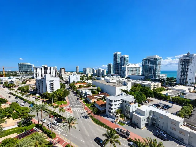 Elevated aerial perspective of a coastal cityscape featuring high-rise buildings, urban thoroughfares, tropical palm tree landscaping, and coastal waters on the horizon