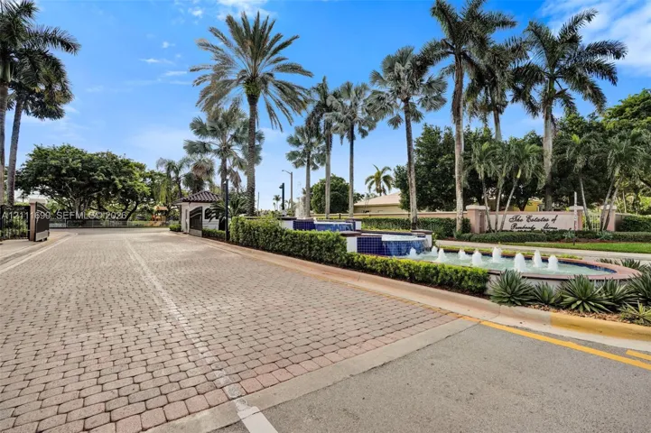 Gated community entrance featuring a paver brick roadway, multi-tiered stone fountain, and mature palm landscaping