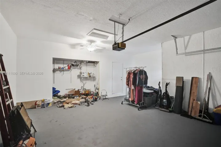 Garage interior featuring a painted concrete floor, ceiling-mounted automatic door opener, ceiling fan with light fixture, and recessed storage alcove