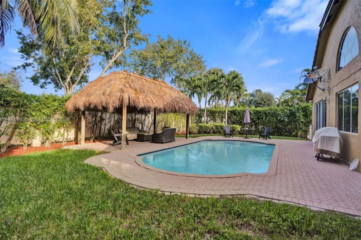In-ground swimming pool with paver decking, thatched-roof tiki hut, and privacy-hedged backyard