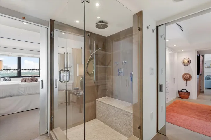 Walk-in glass-enclosed shower featuring a built-in stone bench, overhead rainfall showerhead, handheld sprayer, and neutral tile wall cladding