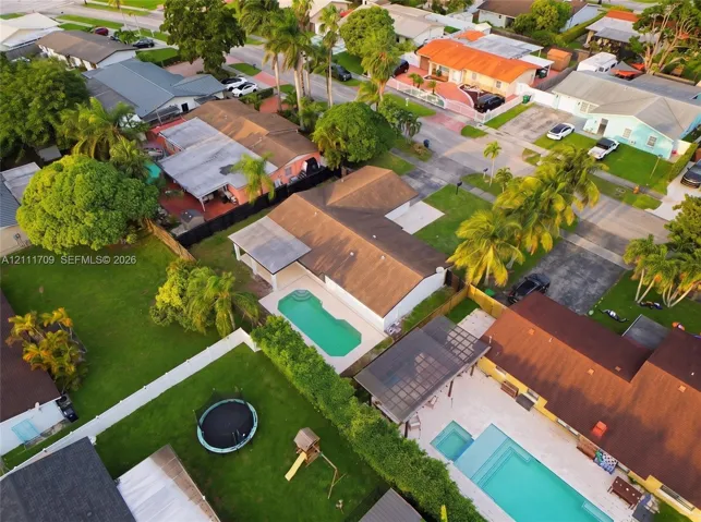 Aerial perspective of a single-story residence featuring a shingled roof, a covered patio, and an in-ground swimming pool situated on a fenced lot with mature landscaping