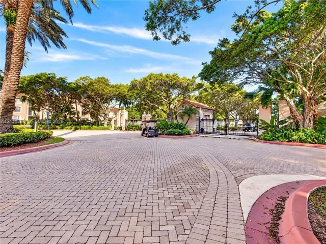 Gated community entrance featuring decorative paver driveway, mature landscaping, and established trees