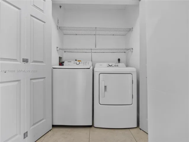 Laundry area featuring a top-load washer, a front-load dryer, dual wire shelving units, and tile flooring, enclosed by white bi-fold doors