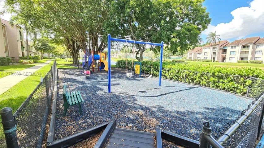 Community playground featuring a blue metal swing set, slide apparatus, and rubberized ground cover, enclosed by chain-link fencing and shaded by mature trees