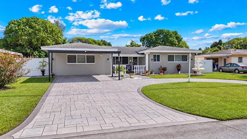 Single-story exterior featuring a paver driveway, covered carport, manicured lawn, tiered water fountain, and white balustrade railing