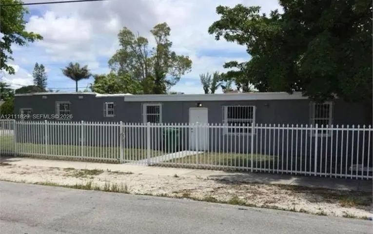 Single-story exterior featuring a gray stucco facade, flat roofline, metal window security bars, and a full-perimeter white metal picket fence