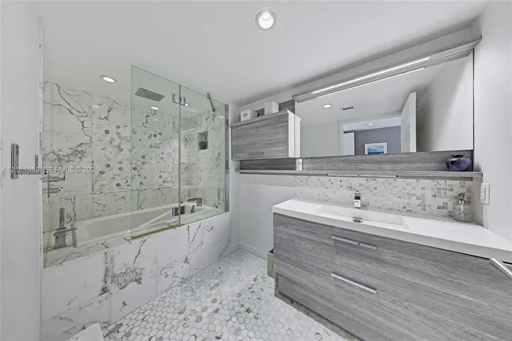 Bathroom featuring a wood-finish floating vanity with an integrated sink, a glass-enclosed soaking tub with stone-tiling, a wall-to-wall mirror, and hexagonal tile flooring