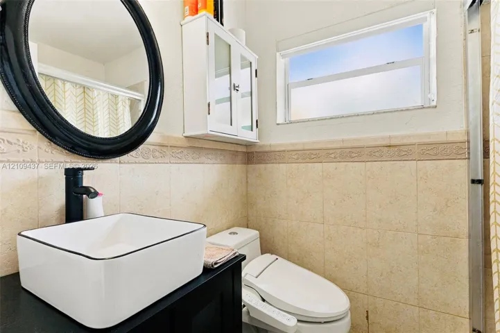 Rectangular vessel sink with matte black faucet, dark vanity cabinet, circular mirror, wall-mounted storage cabinetry, and tiled wall wainscoting