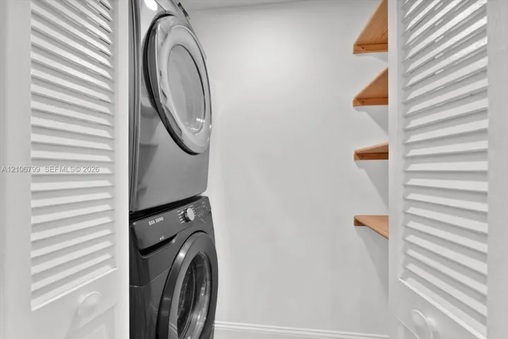 Laundry closet featuring stacked high-capacity appliances, white louvered doors, and built-in wood shelving
