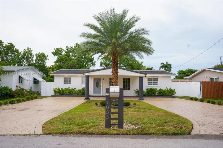 Single-story residence featuring a covered entryway with structural columns, dual paved driveways, a central palm tree, and white privacy fencing
