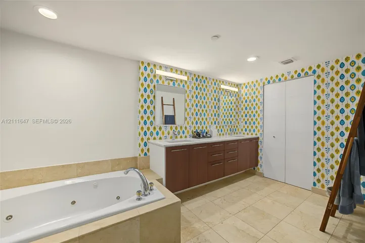 Jetted soaking tub with tile surround, dual-sink vanity with wood-finish cabinetry, stone tile flooring, and integrated wall lighting