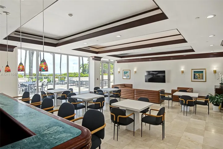 Spacious common area featuring recessed tray ceilings with dark wood trim, tile flooring, built-in stone-top bar, and expansive floor-to-ceiling glass wall panels