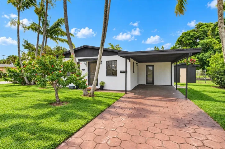 Single-story exterior featuring white stucco siding, a covered carport, hexagonal paver driveway, dark-toned trim, and established palm trees