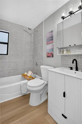 Bathroom featuring a ribbed-detail vanity with a matte black faucet, wood-finish flooring, a tub-shower combination with full-height stone-look tile surround, and a tri-light vanity fixture