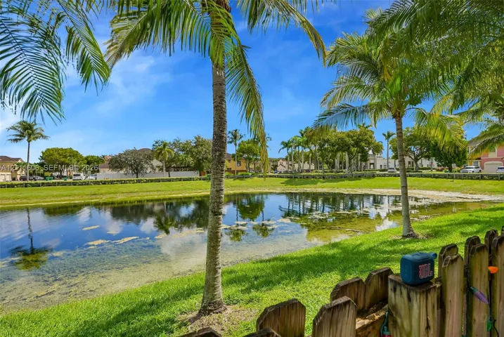 Waterfront lot featuring a pond, manicured lawn, and mature palm trees enclosed by a wooden picket fence
