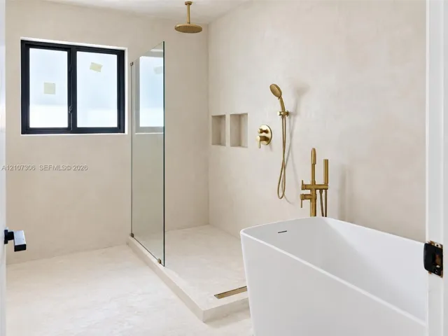 Freestanding soaking tub with brass fixtures, glass-enclosed walk-in shower, dual recessed wall niches, and matte-finish plaster wall treatment