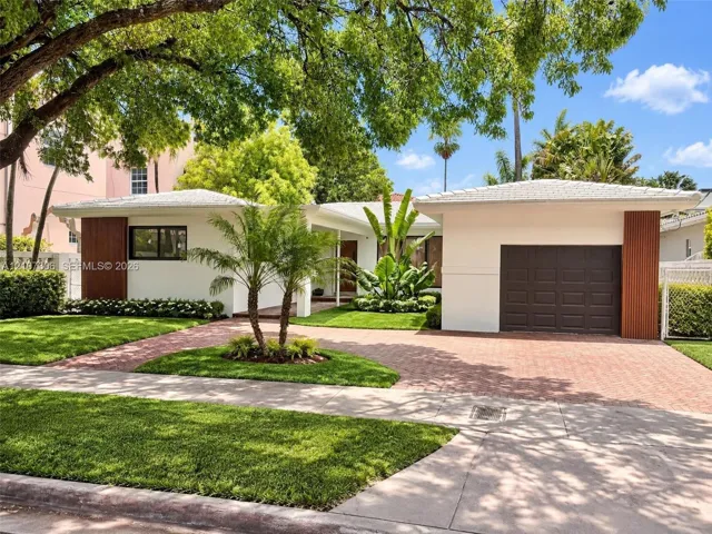 Single-story residence featuring a crisp white exterior with vertical wood-accented columns, a paved brick driveway, a dedicated garage, and mature landscaping