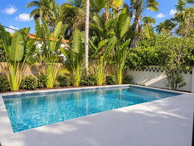 In-ground swimming pool with mosaic tile border, light-toned stone-finish patio, decorative masonry screen wall, and perimeter tropical landscaping